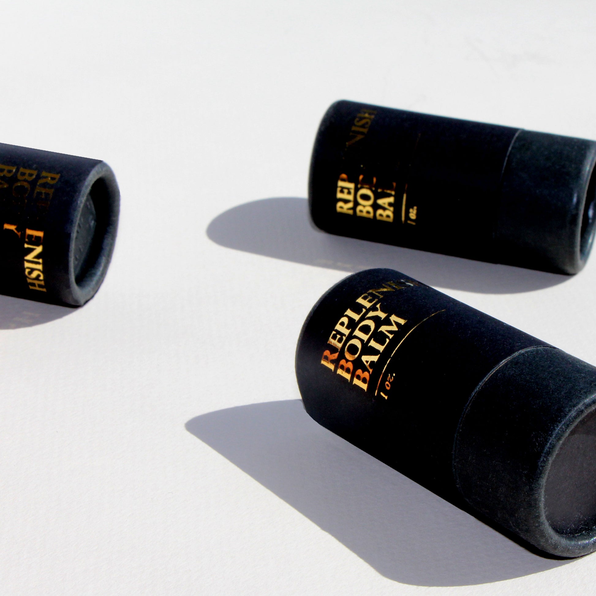 a closeup shot of three black tubes labeled in gold: replenish body balm