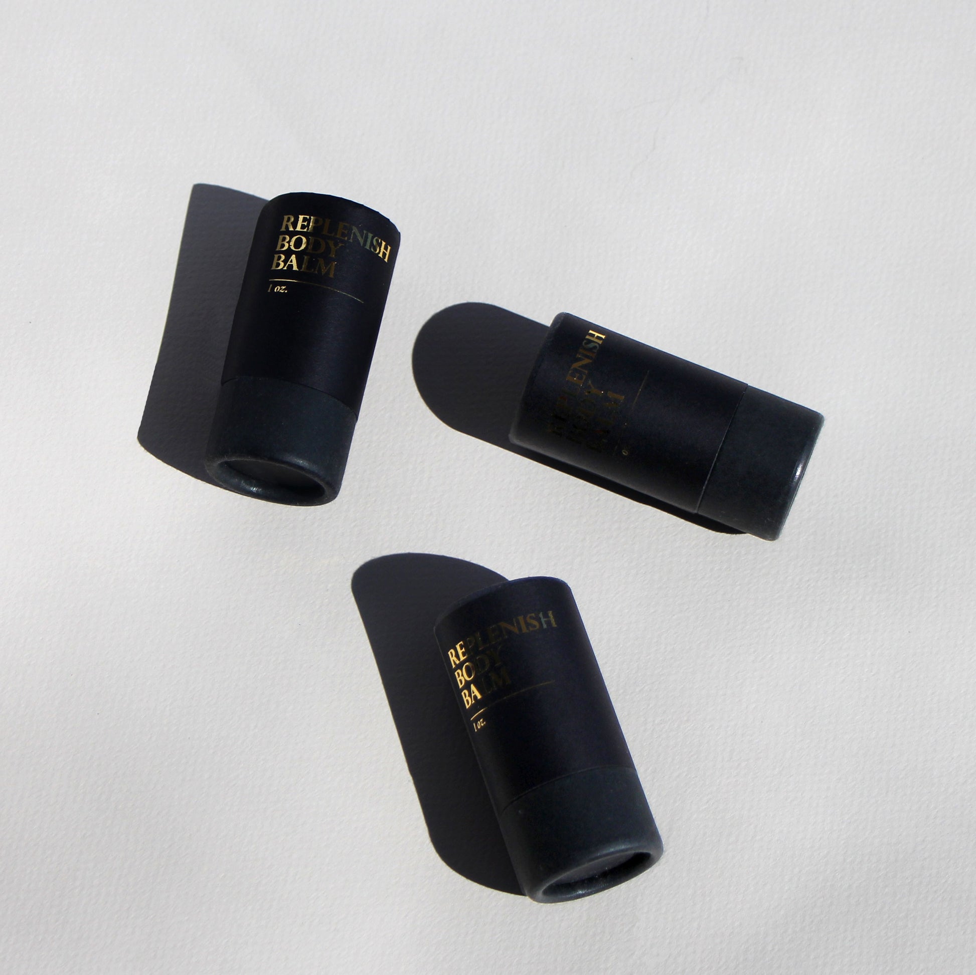 overhead shot of three black cardboard tubes labeled in gold: replenish body balm
