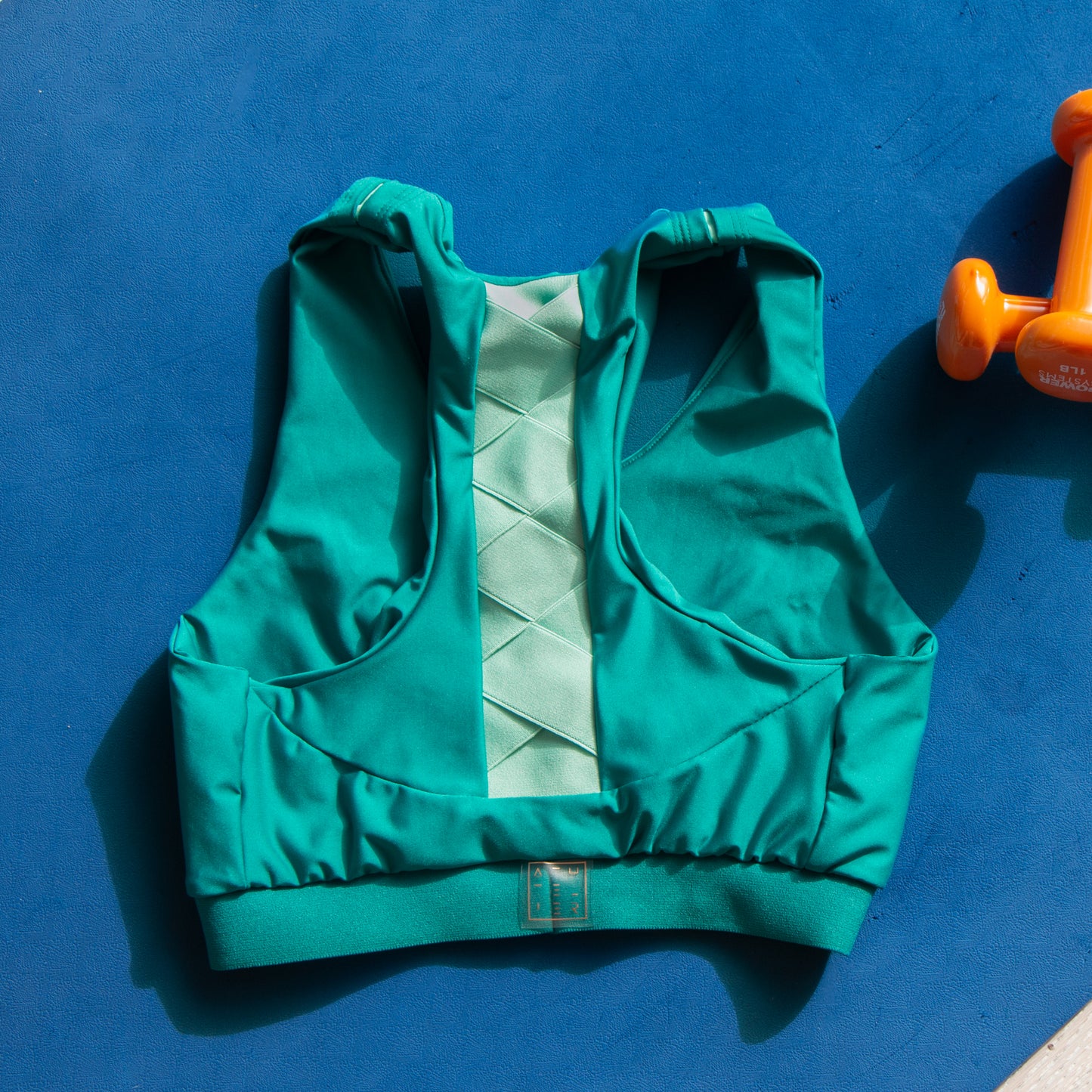 a photo of a green and jade sports bra laying on a blue yoga mat with orange dumbells beside it